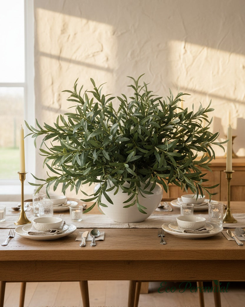 Eco Perennial artificial olive branches with dense green leaves overflowing a wide white ceramic bowl on a rustic oak farmhouse dining table with golden candlelight