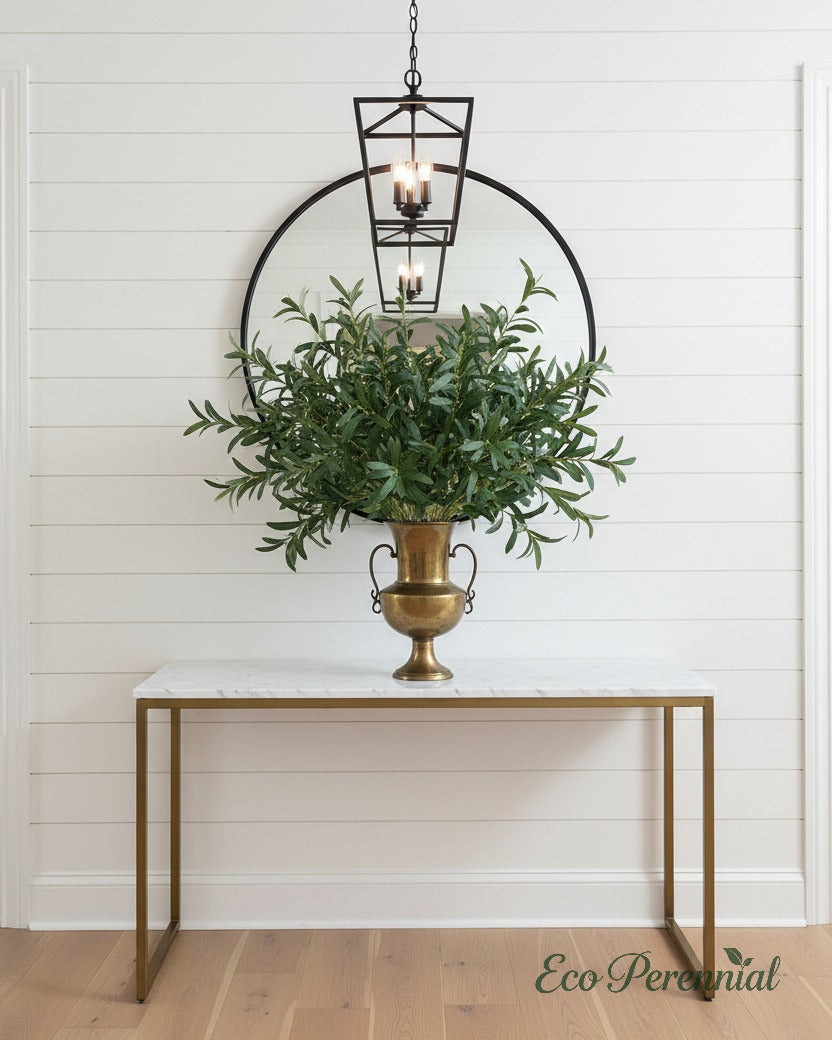 Eco Perennial faux olive branches with abundant green leaves in an antique brass geometric vase on a marble entryway console table with round black-framed mirror